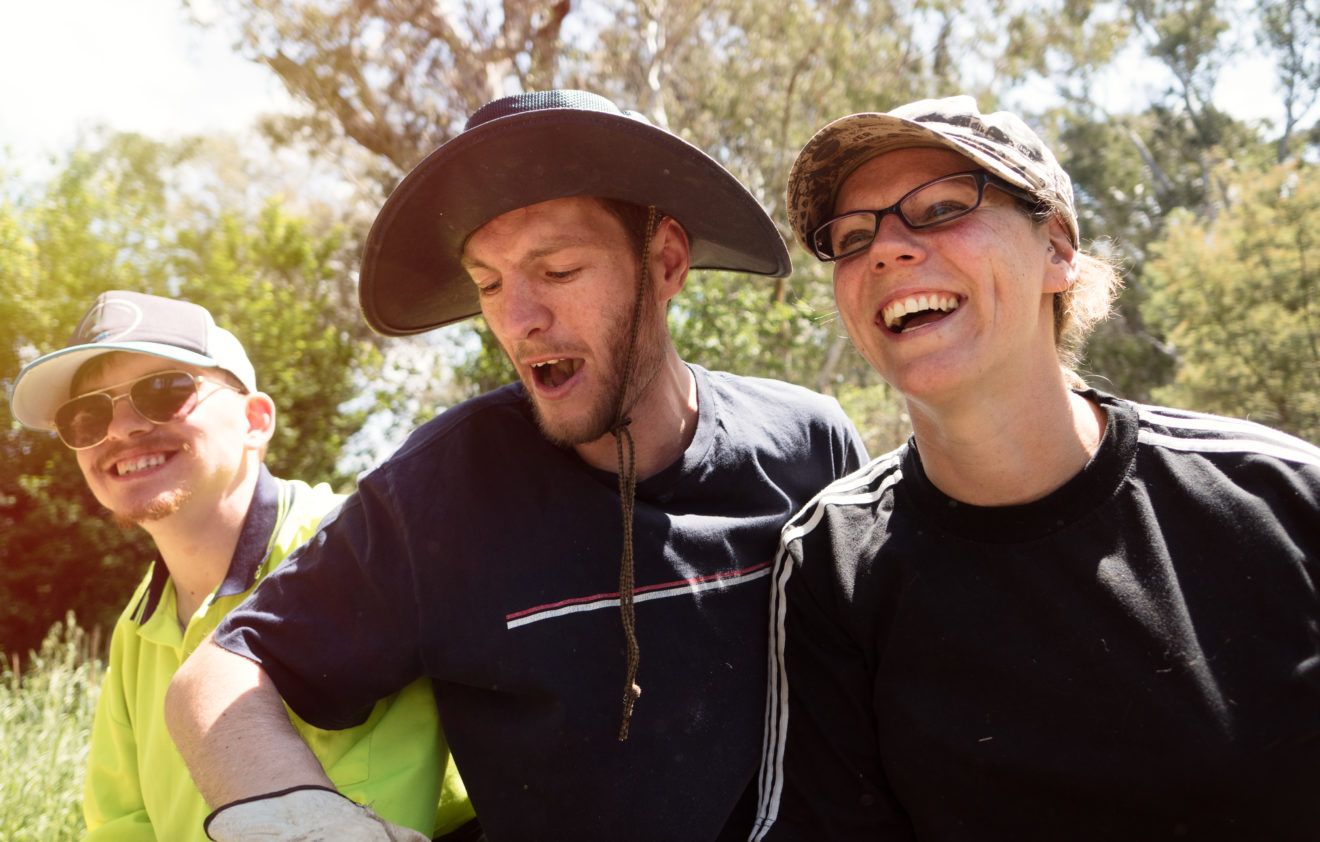 Three young people sitting outside in the sunshine, all wearing hats and having a laugh. Bushland can be seen in the background. 