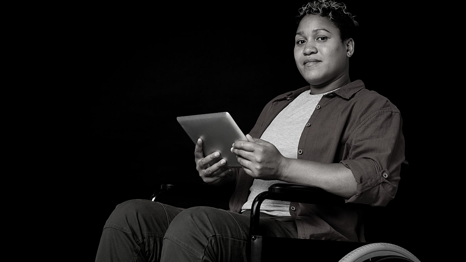A person holding an iPad while looking directly at the camera with a somewhat serious look on their face, and is in a wheelchair. The background behind is pure black.