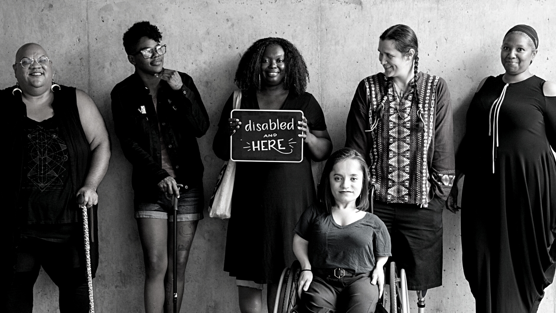 Six people of colour and with disability smile and pose in front of a concrete wall. Five people stand in the back, with the woman in the center holding up a chalkboard sign reading "disabled and here." A person in a wheelchair sits in front.