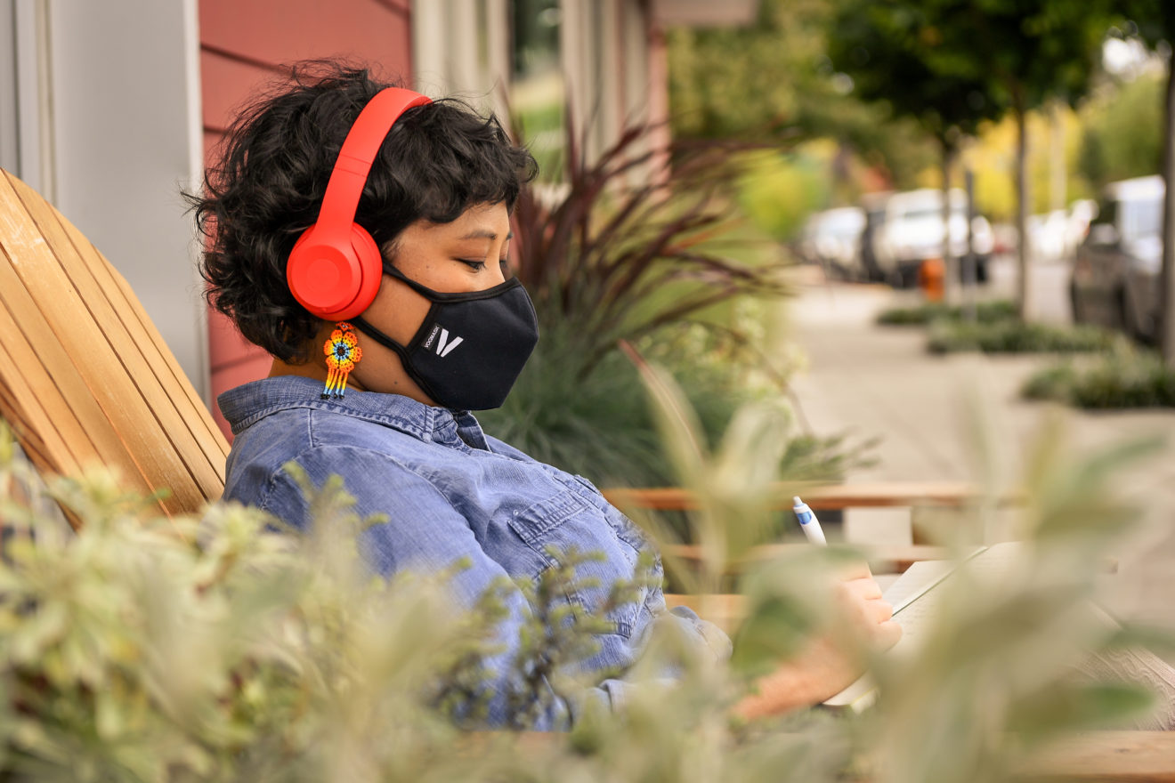 Person viewed from side on and from behind some plants, they are wearing a face mask and headphones and sitting outside a cafe on a deck chair and writing in a notebook. 