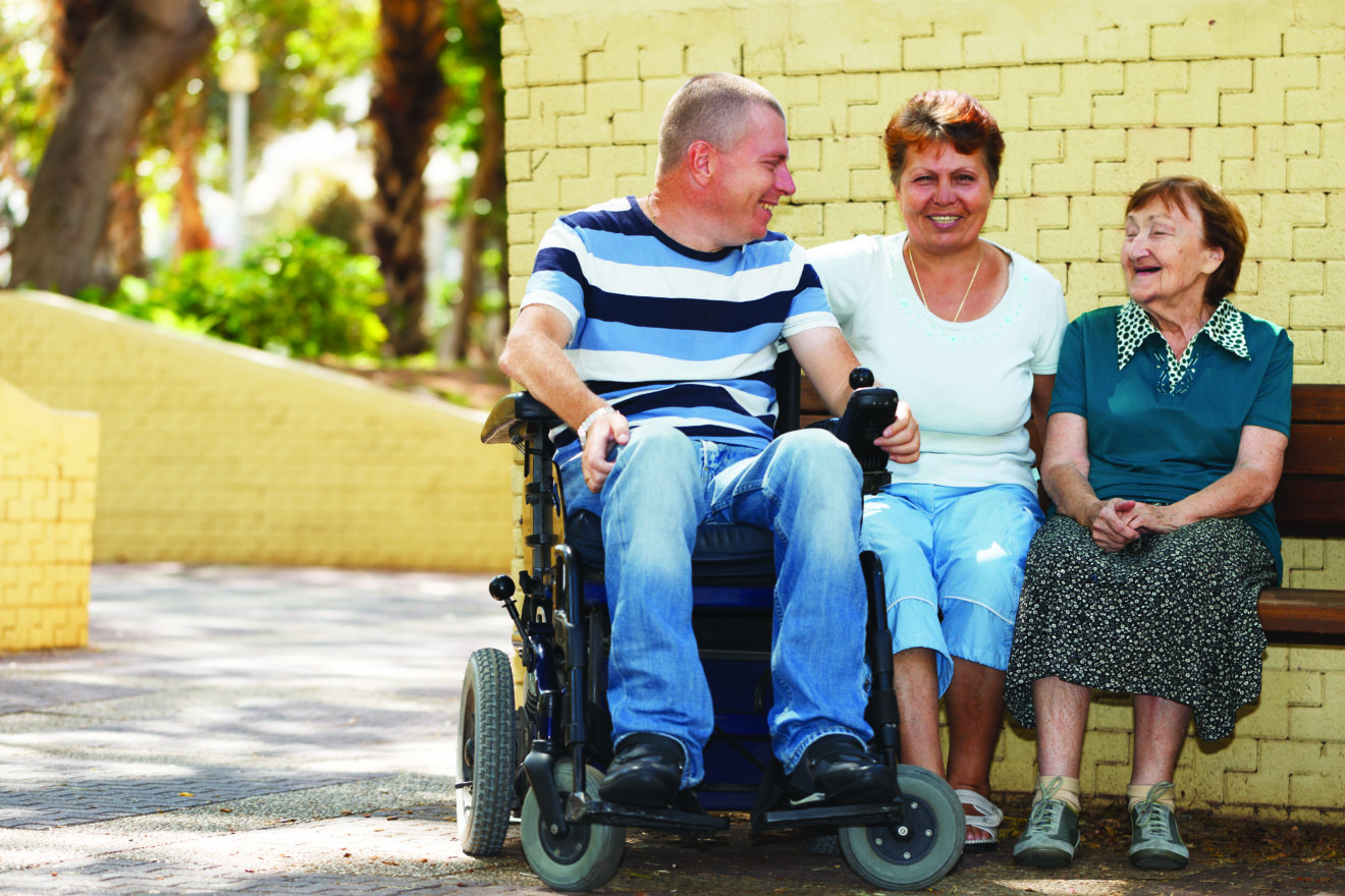 Three people enjoying sitting outside and chatting on a sunny day, two are sitting on the end of a bench seat directly in front of a brick wall, and a third is sitting in a wheelchair right next to them. 