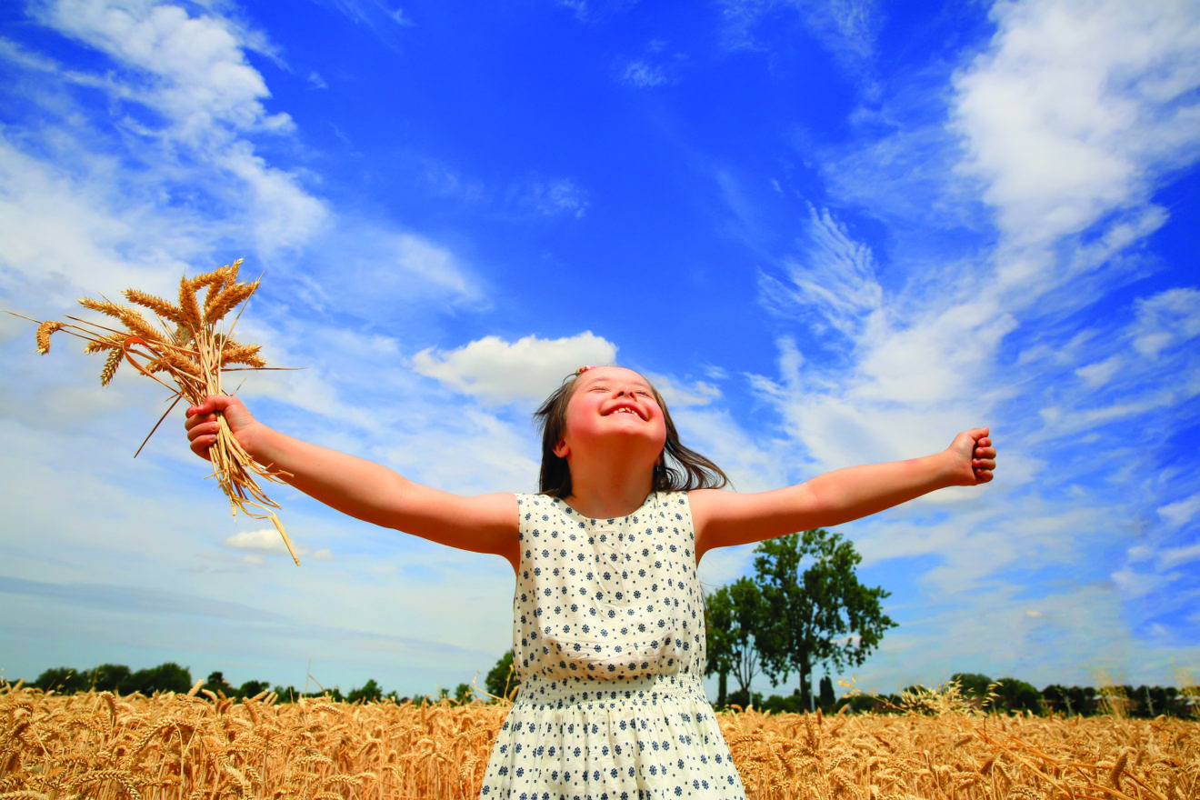 Young girl standing in a wheat field on a bright sunny day, with her arms outstretched on either side and holding a bunch of wheat stalks in one hand, while she looks up at the blue sky dotted with clouds. Her eyes are closed and she is smiling ecstatically.