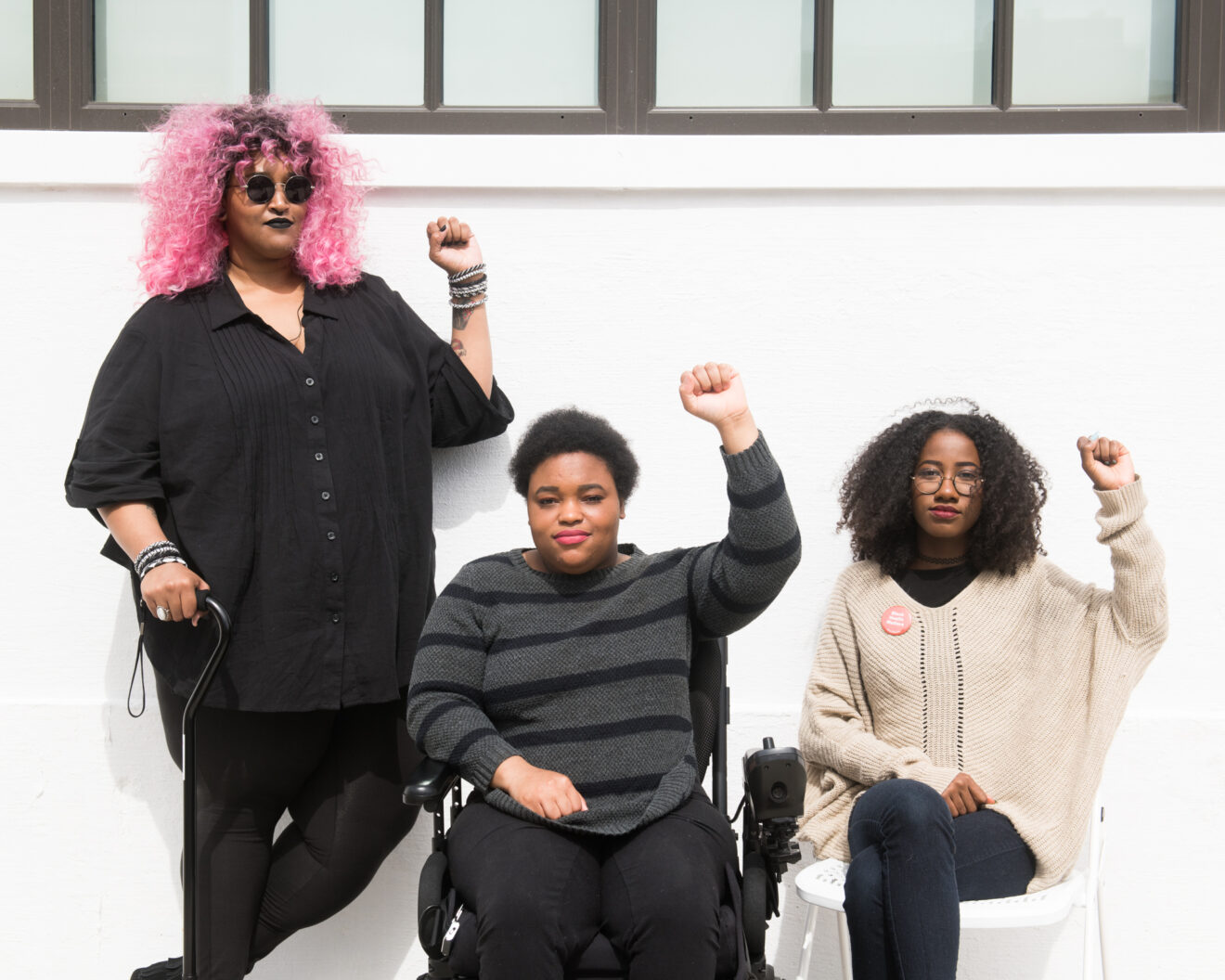 Three people, one holding a cane, one in a power wheelchair, and on a chair raising their fists in front of a white wall.