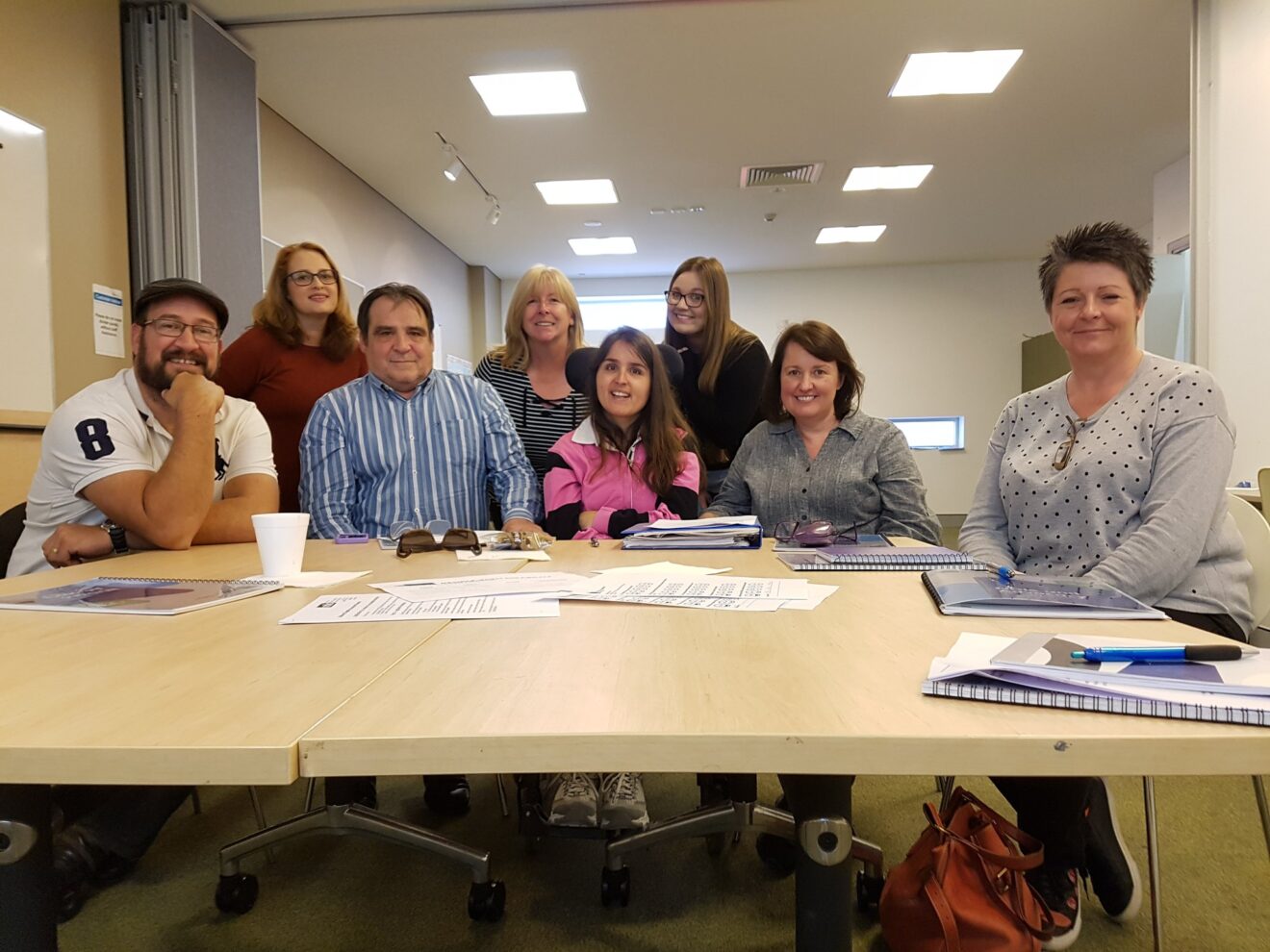 Eight people attending PWDA's NDIS Appeals Training sitting around a desk looking at the camera smiling.