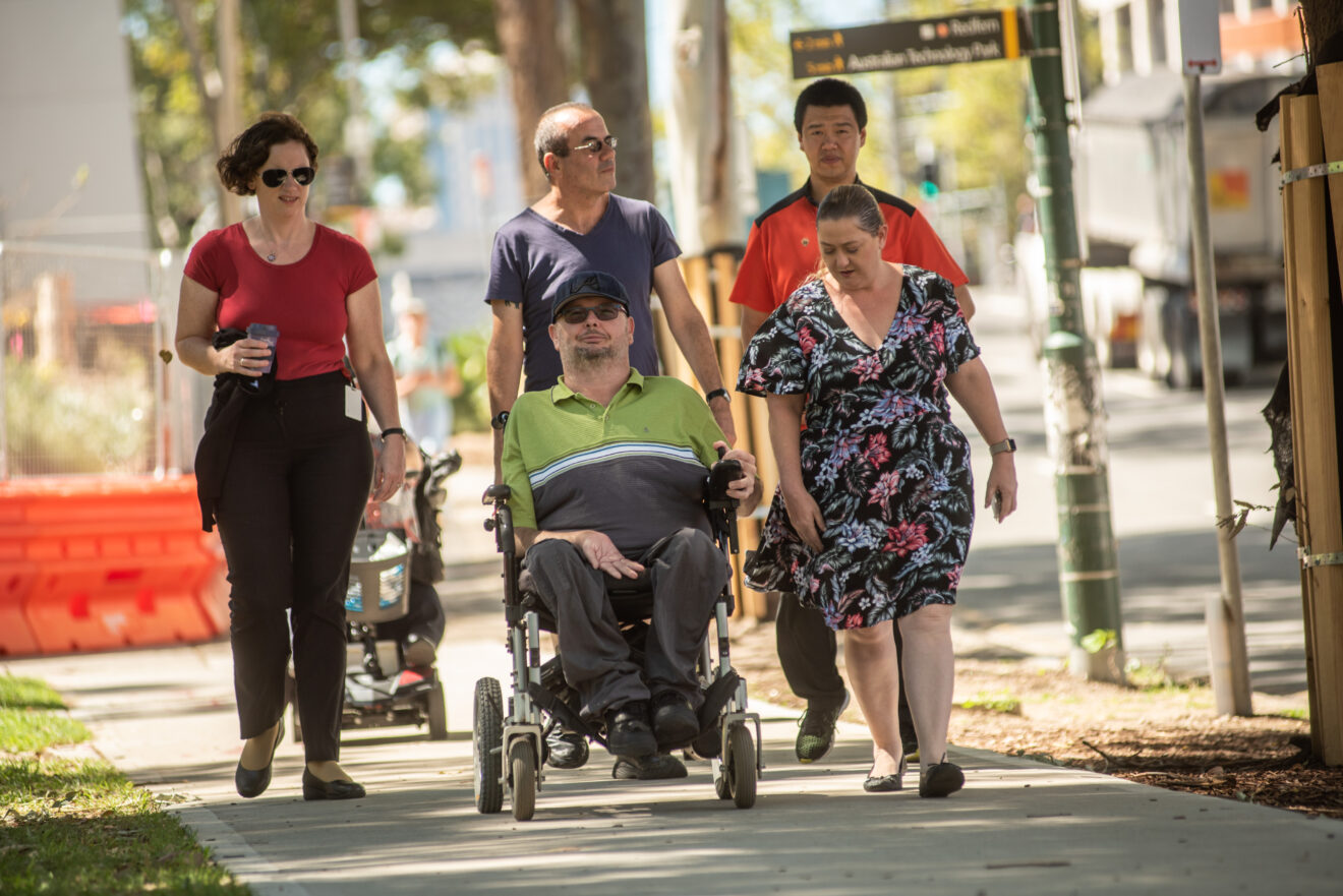 A group of people walking down and some using a wheelchair a footpath, talking to each other.