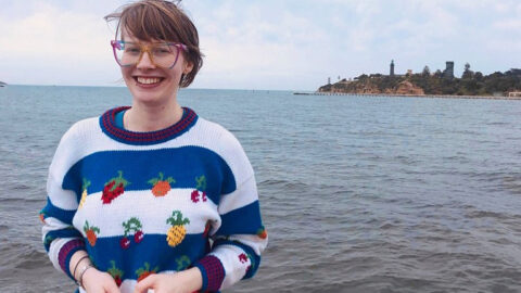 [Photograph of Alex Creece, a white woman with short hair, large colourful glasses, a knitted jumper with a fruit pattern, smiles at the camera. Behind her is the ocean, a peer, and part of the shore.]