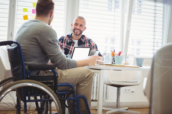 Two people sitting at a table in a sun-drenched office space. The person in the foreground is in a wheelchair and has a laptop on in front of him, and the other person is facing him and smiling.