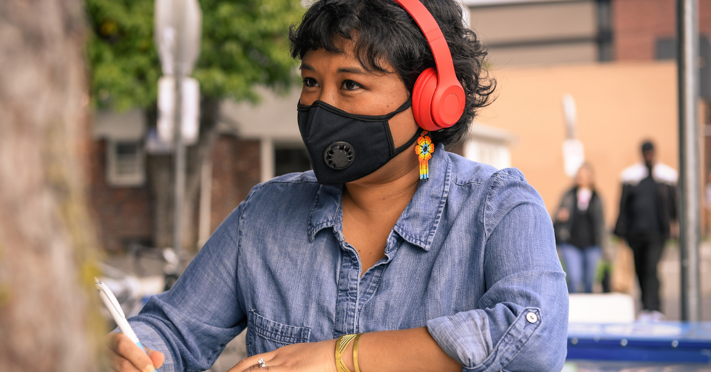 Close-up of a Filipinx woman with a filtering face mask, sitting at a table with notebook and pen. She has colourful flower earrings and headphones on while looking into the distance.