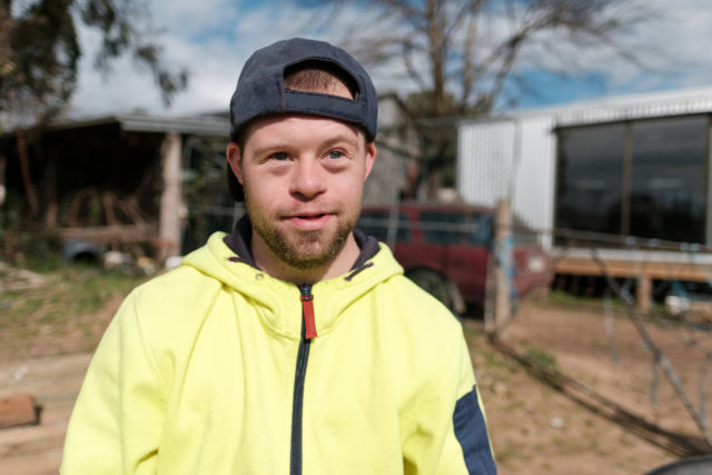 Close up of a young man on a farm, smiling slightly at the camera. He is wearing a cap on backwards and a yellow hoodie. Farm buildings can be seen in the background.