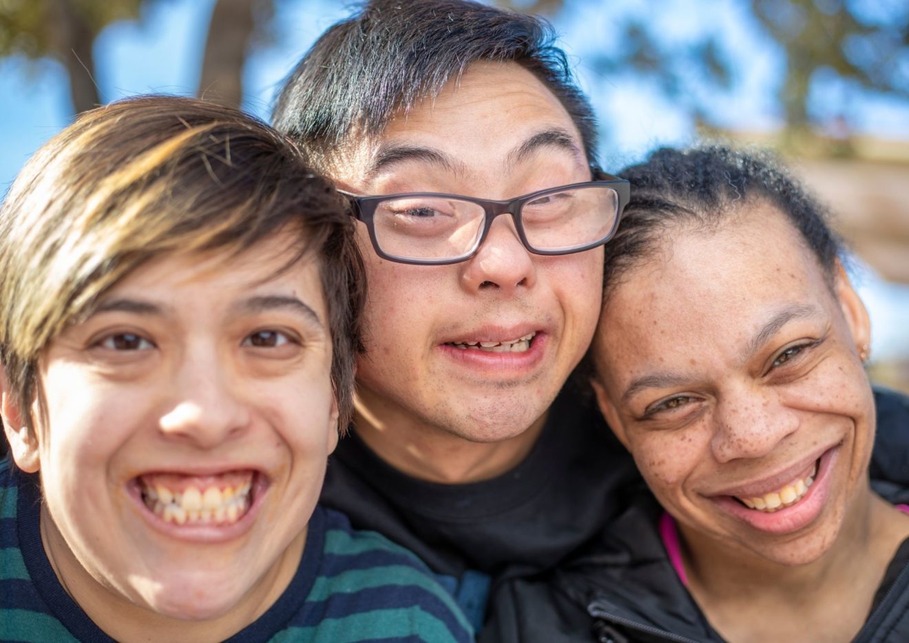 Close up of three young people with disability huddled closely together and posing and smiling for the camera