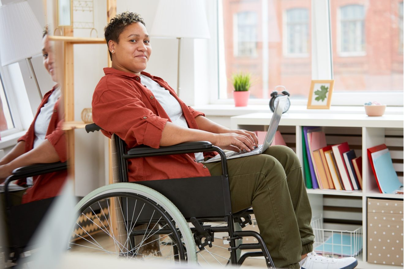 A person viewed from side on, sitting in front of a large window in a wheelchair and smiling. There is a laptop resting on their lap, and they are wearing khaki pants, white t-shirt and a re shirt over the top.