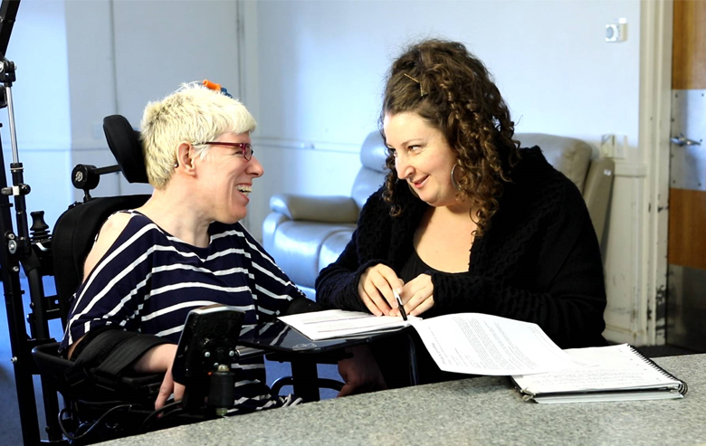 A disability support worker and their client, who is in a wheelchair, sitting in front of a table in a room. They are smiling at each other, and the support worker is holding a pen which is poised over a section of text on the multi-page report in front of them.