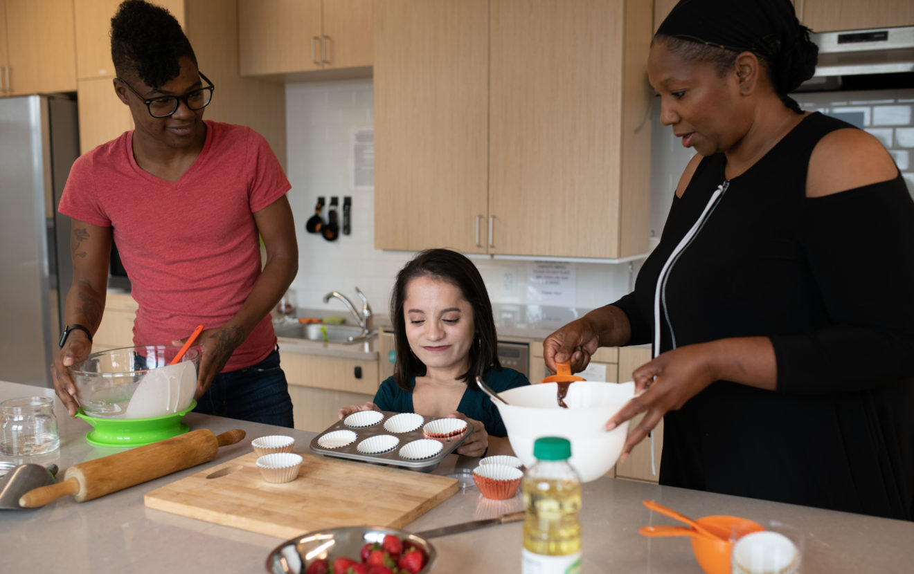 Three people baking in a kitchen, person on left holds a mixing bowl,  person in the middle prepares a muffin pan and person on the right measures out a chocolate mix.