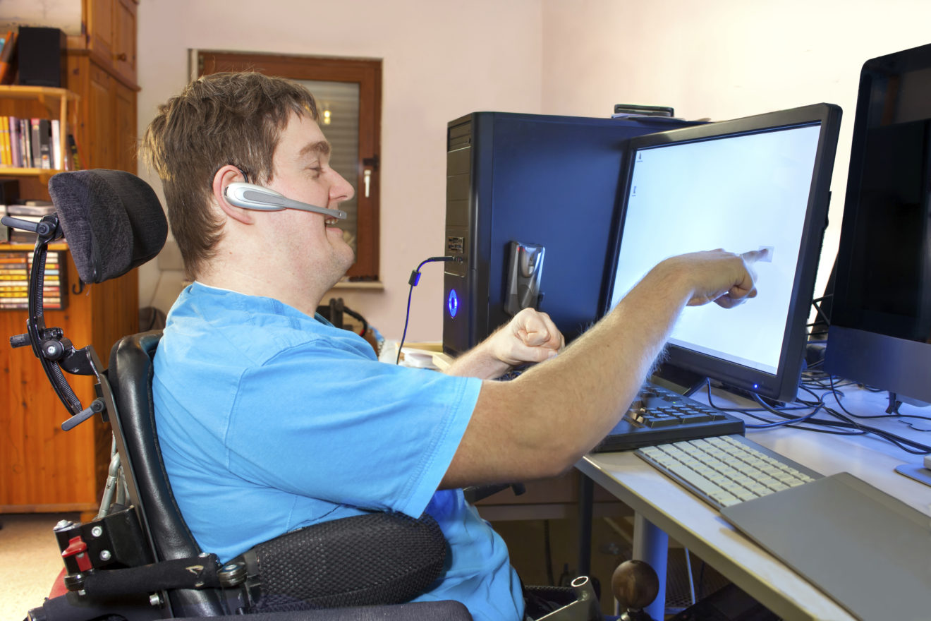 Man with disability sitting in multifunctional wheelchair, using a computer with a wireless headset, reaching out to touch the touch screen.