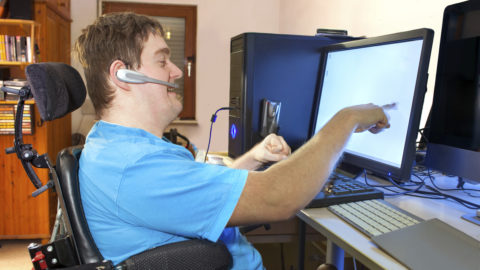 Man with disability sitting in multifunctional wheelchair, using a computer with a wireless headset, reaching out to touch the touch screen.