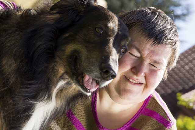 a woman with intellectual disability smiles in sunlight. A dog is to her left