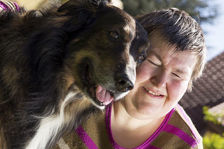 [IMAGE a woman with intellectual disability smiles in sunlight. A dog is to her left]