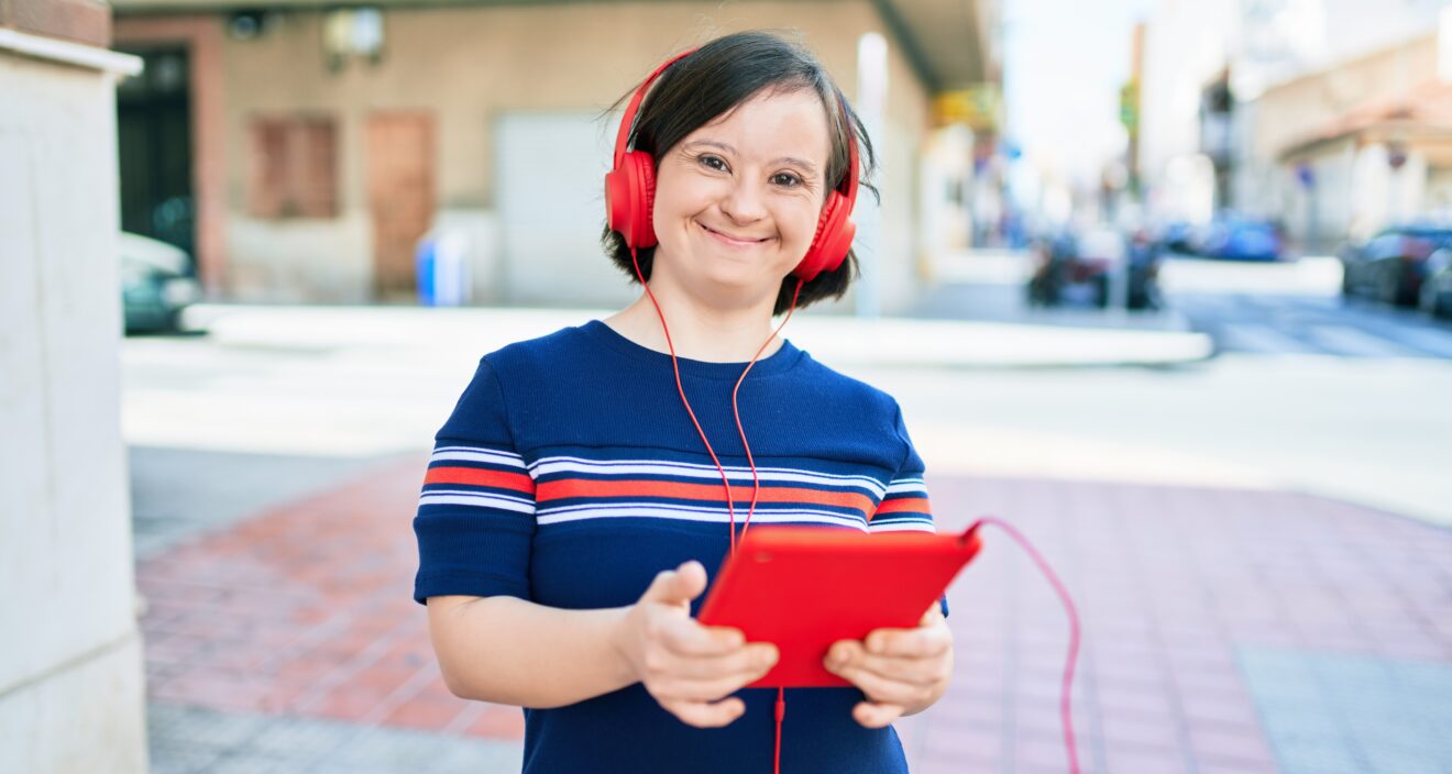 Person smiling, standing on a suburban street on a sunny day using touchpad device and wearing headphones.