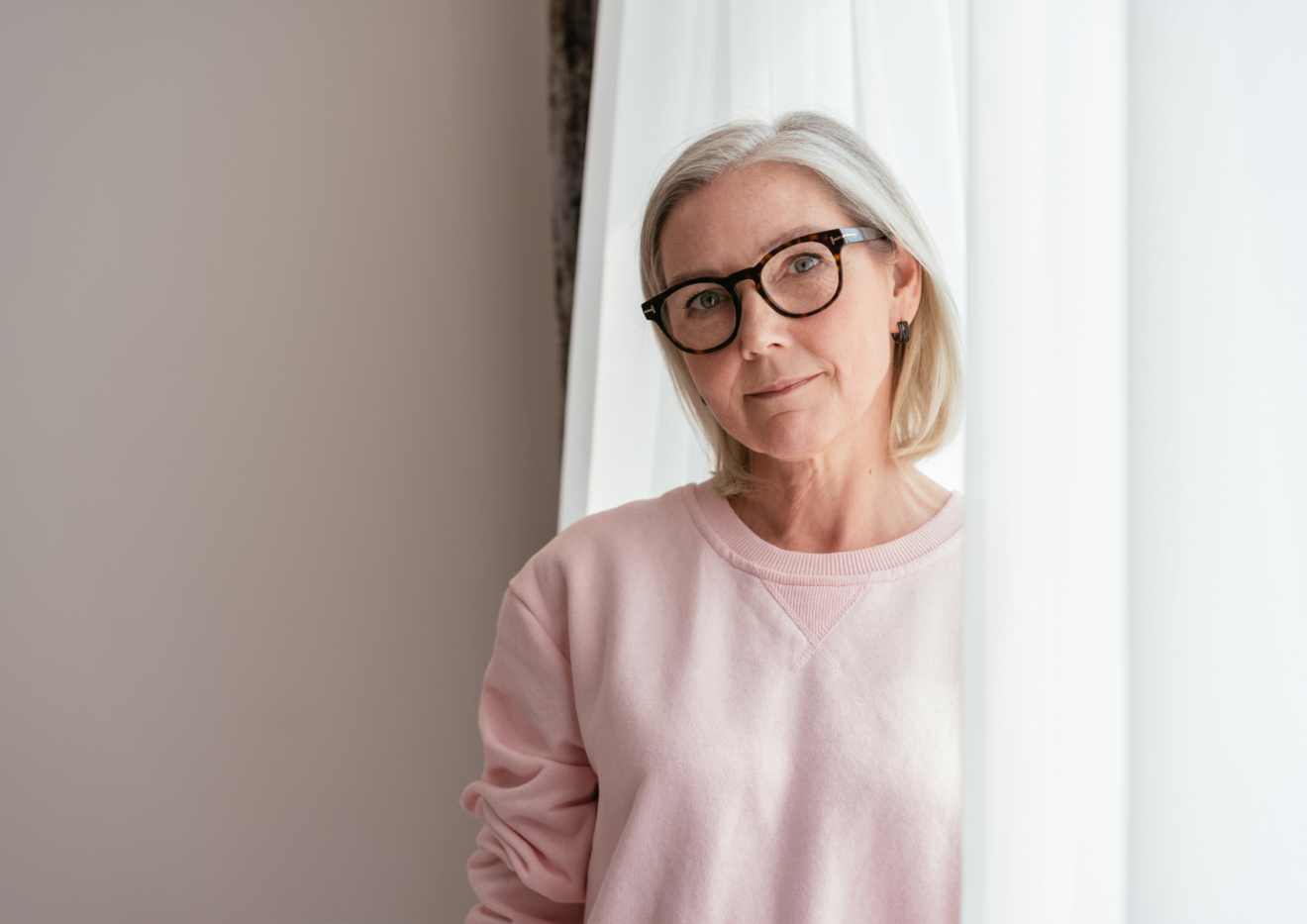 Woman with light grey hair and wearing glasses and pink jumper standing partially in the curtains next to a window