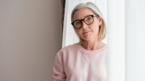 Elderly Woman in Pink Sweater Wearing Eyeglasses - People with Disability Australia Woman with light grey hair and wearing glasses and pink jumper standing partially in the curtains next to a window