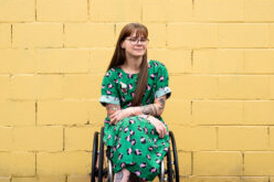 Nicole Lee is positioned in front of a yellow brick wall, she has long brown hair, wearing glasses and a printed green dress and is sitting in a wheelchair.