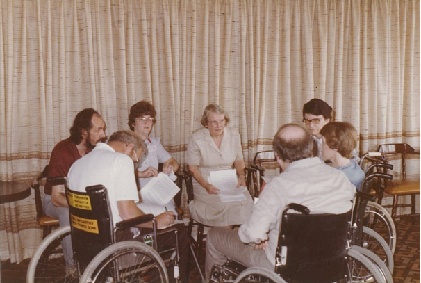 Seven people, mostly wheelchair users, sitting in a close circle in discussion and holding paperwork. The photo was taken in the 1980’s.