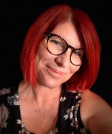 Close up of Samantha Connor who has her head tilted to the side and smiling. She has bright red hair and is wearing glasses and a floral top