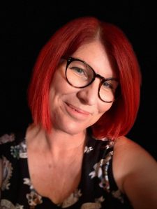 Close up of Samantha Connor who has her head tilted to the side and smiling. She has bright red hair and is wearing glasses and a floral top