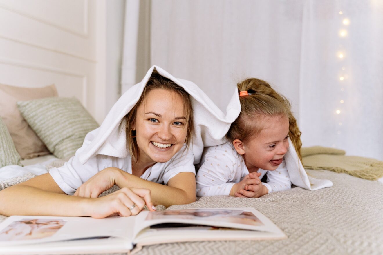 A mother, with an open book in front of her, and daughter looking away, laying on their stomachs on a bed with a towel playfully draped over their heads 