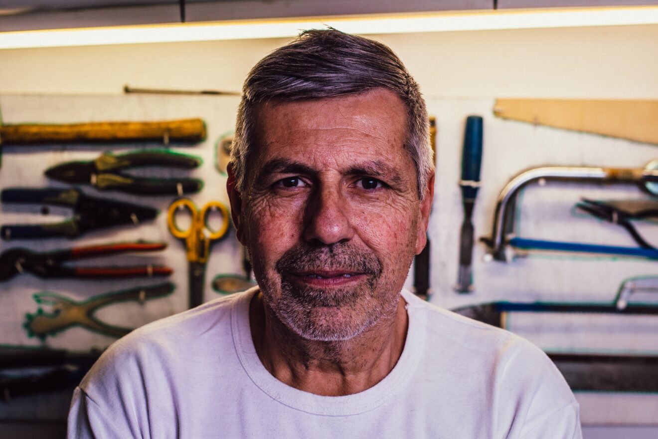 Close up of a man with short grey hair who is looking intently into the camera, he is wearing a white t-shirt and tools can be seen hanging on a wall behind him.