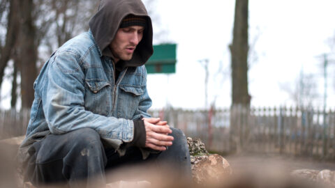 Homeless person sitting outside on a cold grey day, wearing well-worn jacket, long pants, beanie and a hoodie, trying to warm up his hands, and looking sad and alone.