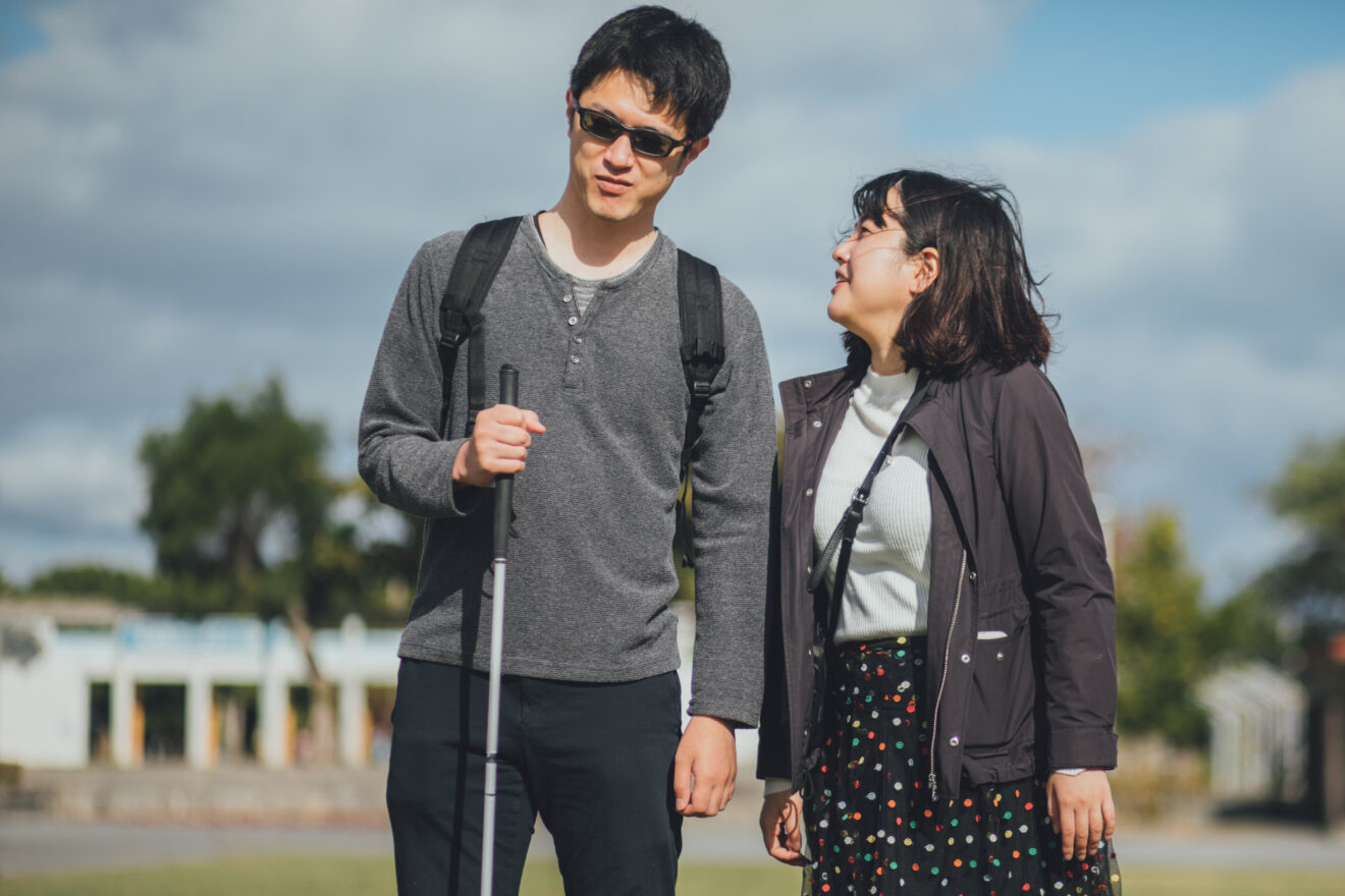 Two people standing closely together outside on a partly cloudy day, the person on the left is holding a white cane and is wearing glasses, the other person is looking up at him and smiling