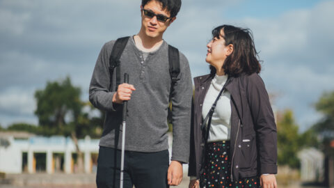 Two people standing closely together outside on a partly cloudy day, the person on the left is holding a white cane and is wearing glasses, the other person is looking up at him and smiling