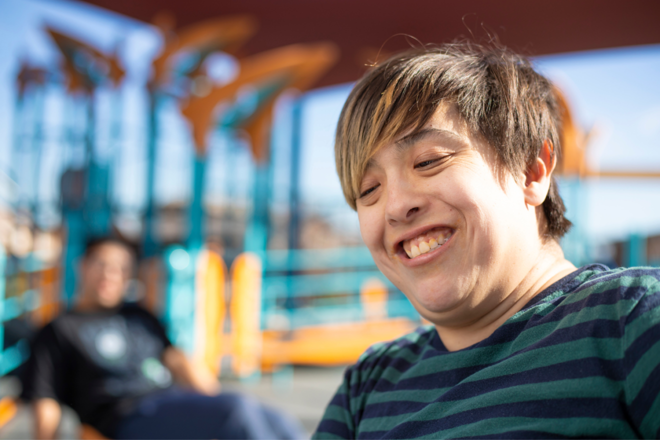 A young person with intellectual disability smiling past the camera. They are outdoors with a friend.