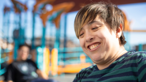 A young person with intellectual disability smiling past the camera. They are outdoors with a friend.