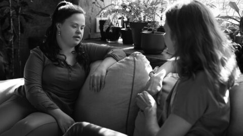 Two women facing each other while sitting casually on a lounge deep in conversation. Directly behind them is a large window with several plants on the window sill.