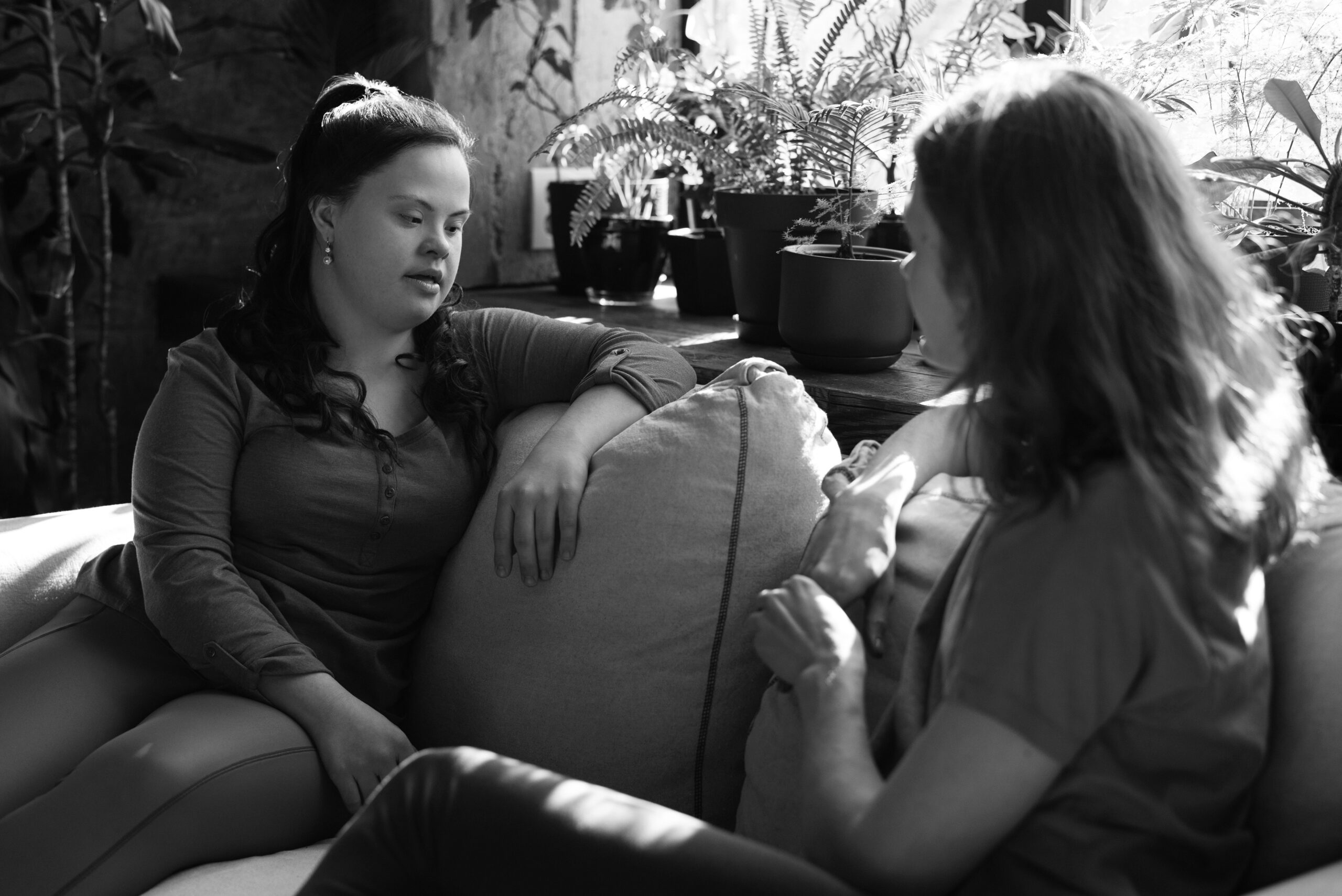 Two women facing each other while sitting casually on a lounge deep in conversation. Directly behind them is a large window with several plants on the window sill.