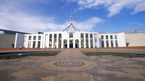 A photograph of Parliament House in Canberra on a bright, sunny day. The picture is taken from across the forecourt, and shows in detail the mosaic on the ground. The artwork is called ‘Possum and Wallaby Dreaming’ by Kumantje Jagamara.
