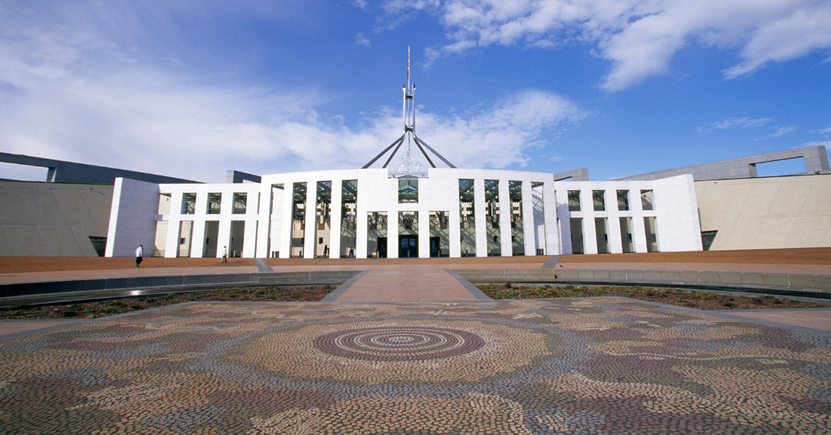A photograph of Parliament House in Canberra on a bright, sunny day. The picture is taken from across the forecourt, and shows in detail the mosaic on the ground. The artwork is called ‘Possum and Wallaby Dreaming’ by Kumantje Jagamara.