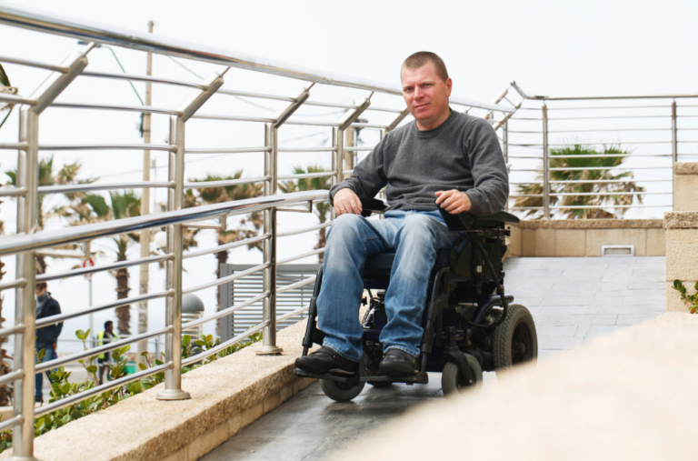 Person, smiling slightly, sitting in a motorised wheelchair on a gently sloping ramp and alongside a long high railing to his left, through which palm trees and water off in the distance can be seen.