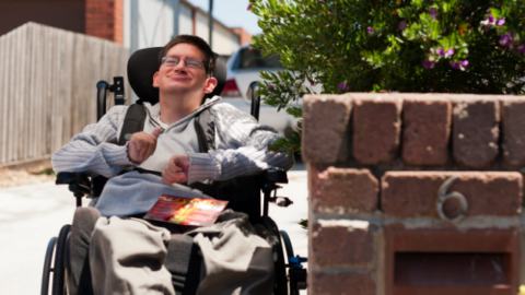 A young male wheelchair user out the front of his house in the driveway after collecting the mail.