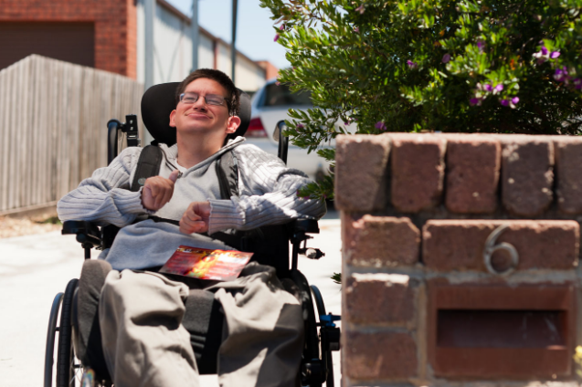 A young male wheelchair user out the front of his house in the driveway after collecting the mail.