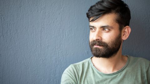 Close up of a young man positioned in front of a grey wall. He is looking off into the distance to the left, and has dark hair, beard and is wearing an olive green t-shirt