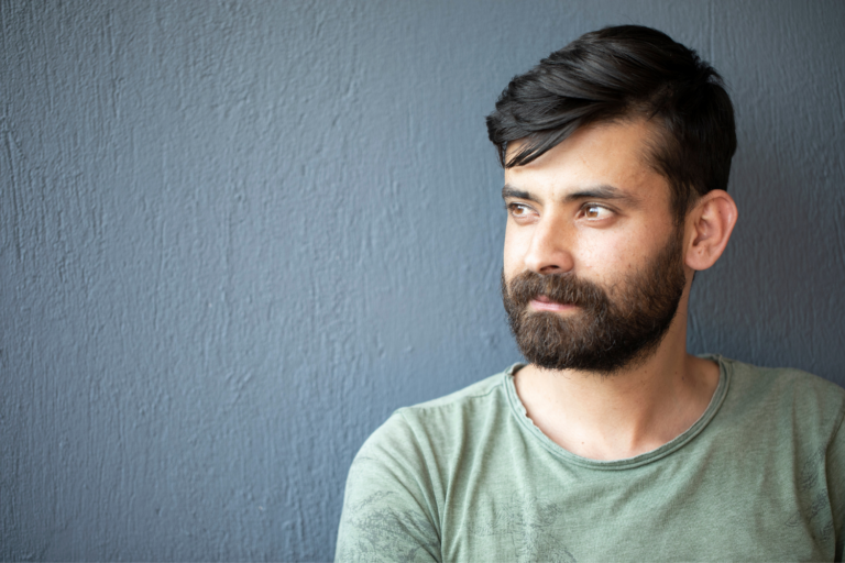 Close up of a young man positioned in front of a grey wall. He is looking off into the distance to the left, and has dark hair, beard and is wearing an olive green t-shirt