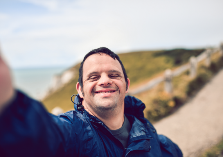 Man taking a selfie photo, as seen from his camera view, and smiling happily. In the background, a path can be seen which continues up the mountain he is on next to the ocean.