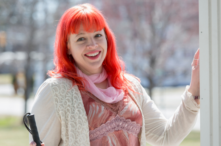Woman standing in a doorway that leads outside on a bright sunny day, with one hand resting on the frame and smiling happily. She has bright pinky red long hair, and is holding a white cane.