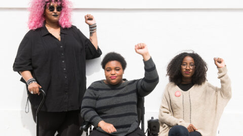 Three people, one holding a cane, one in a power wheelchair, and on a chair raising their fists in front of a white wall.