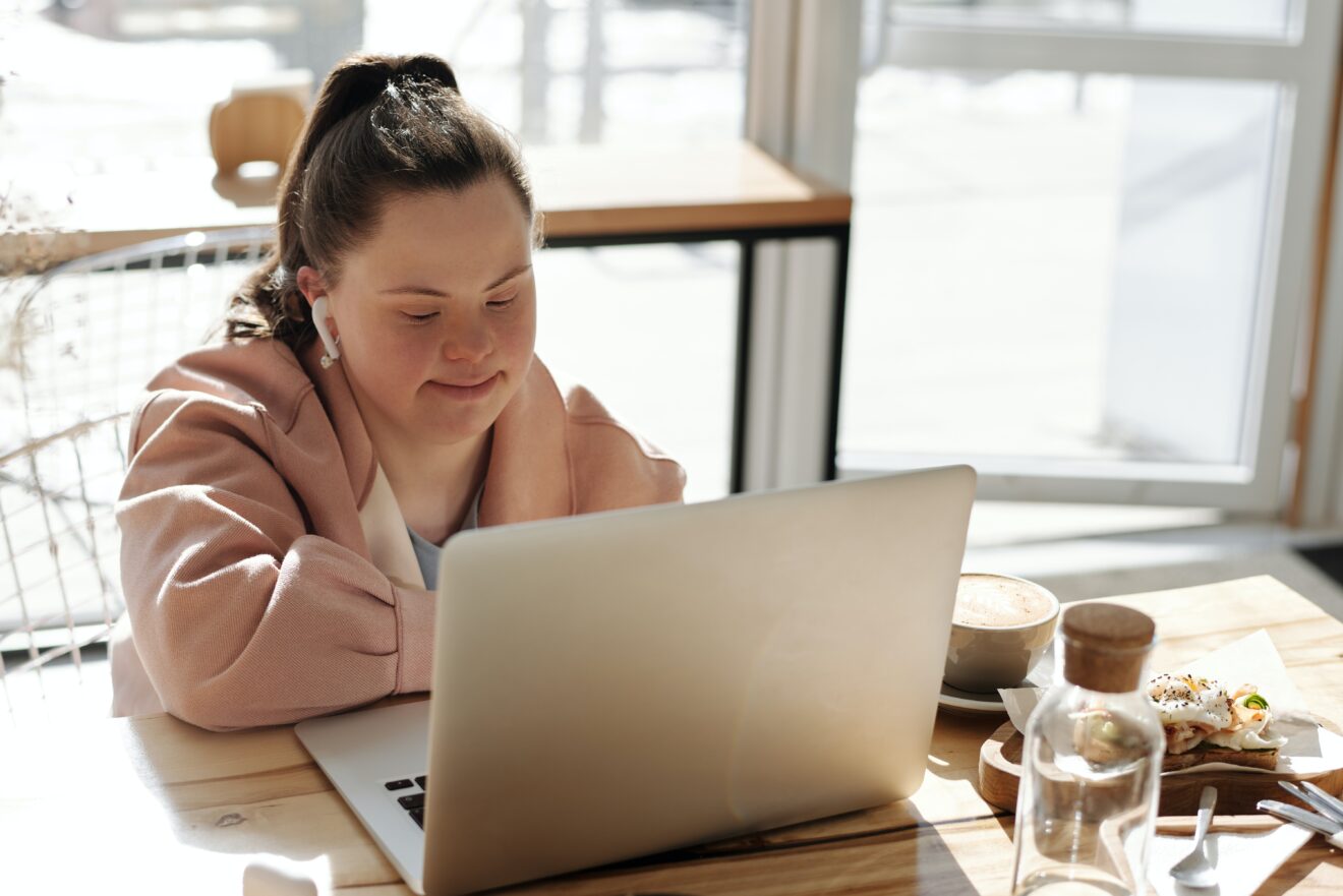 Woman sitting at a cafe table with the sun streaming in behind her, she is looking at an open laptop in front of her, she has earbuds in her ears and coffee and food can be seen on the table beside her.