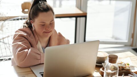 Woman sitting at a cafe table with the sun streaming in behind her, she is looking at an open laptop in front of her, she has earbuds in her ears and coffee and food can be seen on the table beside her.