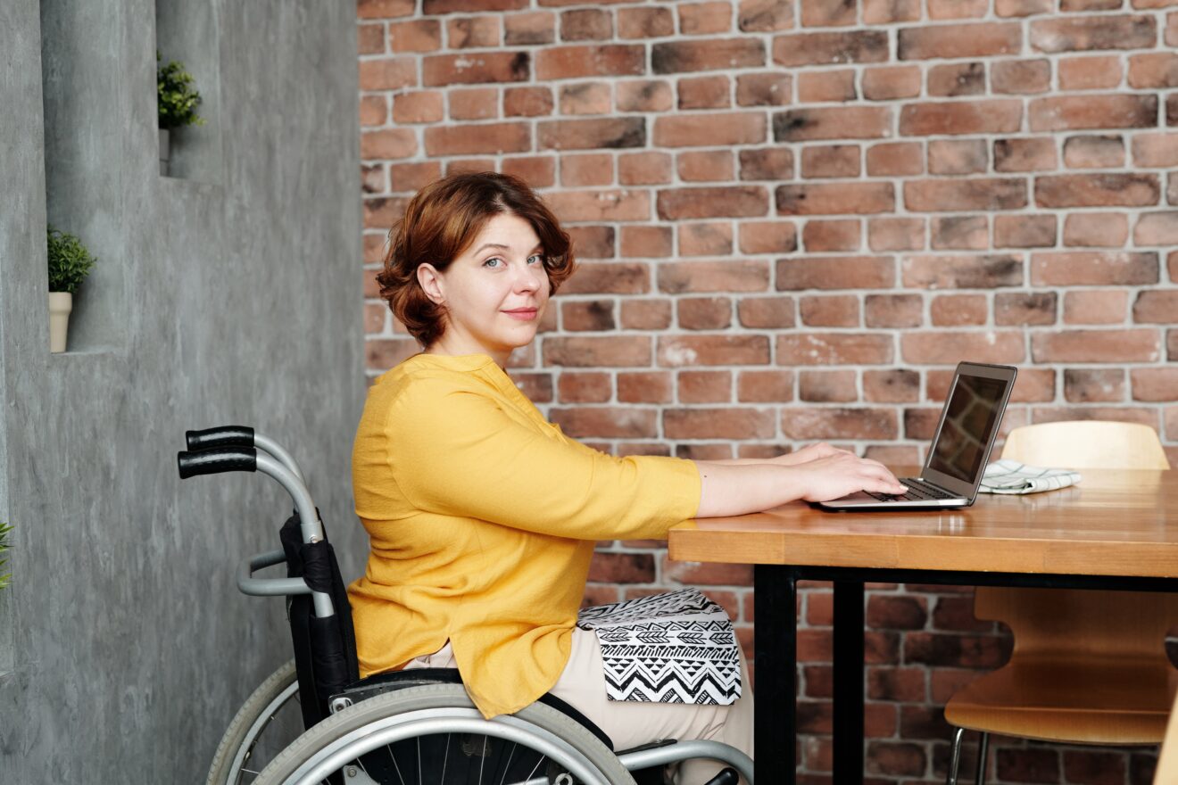 Person viewed side on, sitting in at a table in a wheelchair and her hands are resting on the keyboard of an open laptop in front of them, they are smiling slightly and looking directly at the camera